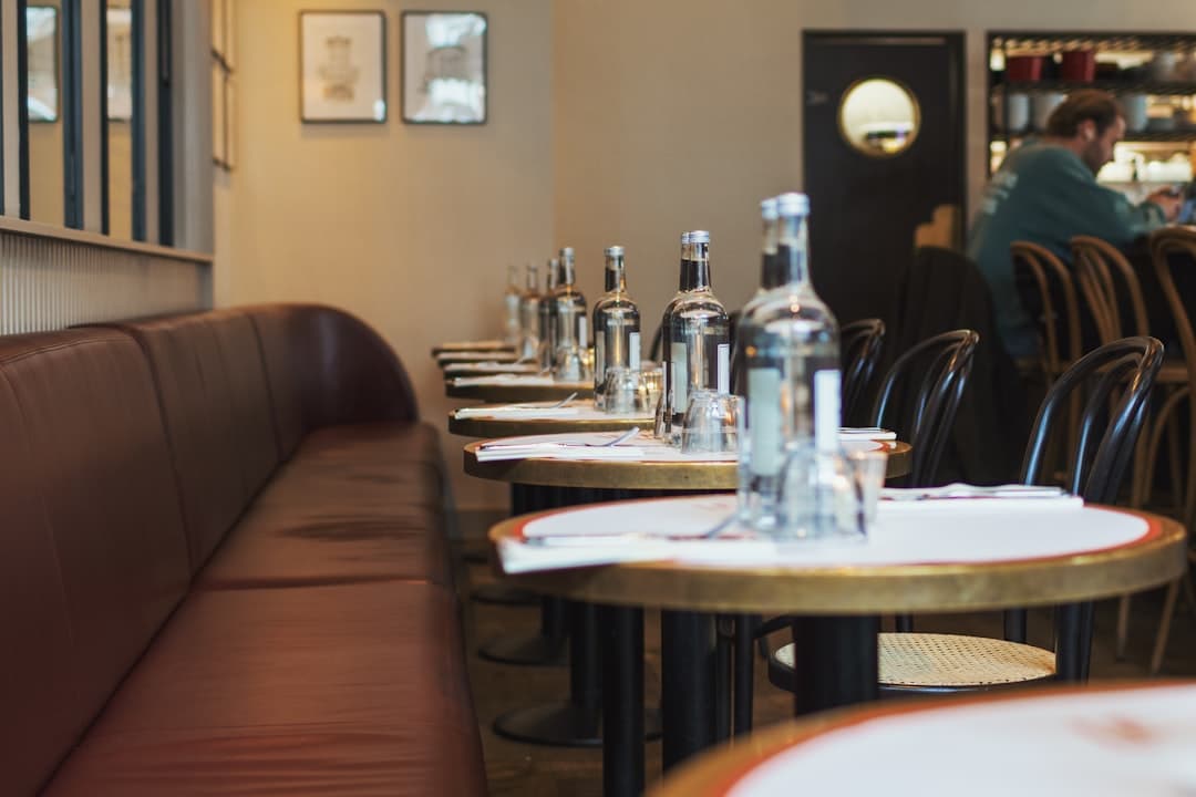 A row of small bistro tables laid with white cloths and water bottles along a leather banquette.