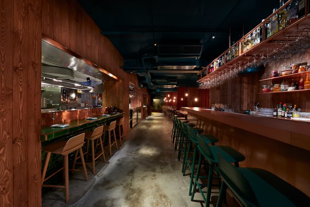 A long wooden bar counter with green stools and bottles lined up on the shelves behind it.