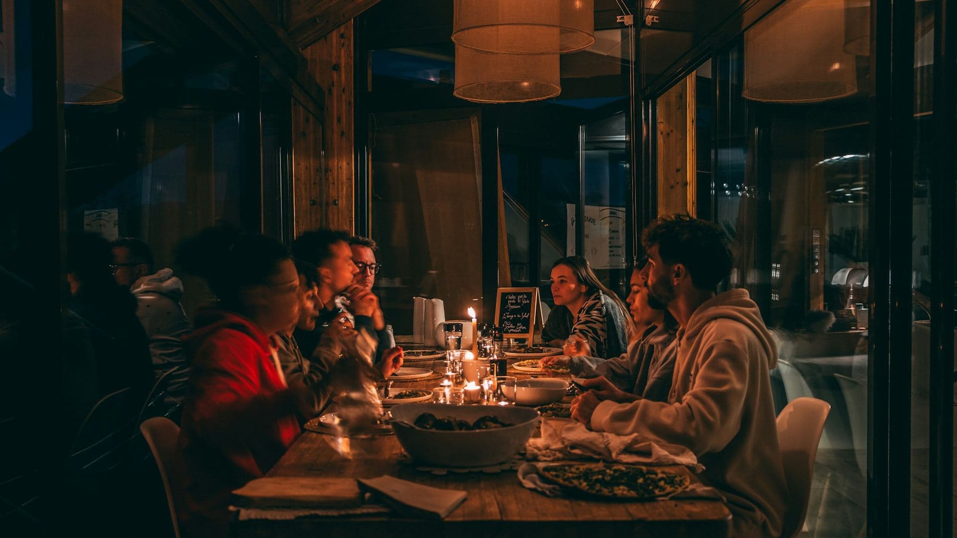 Six people eating together at a long table in a restaurant window, late evening.